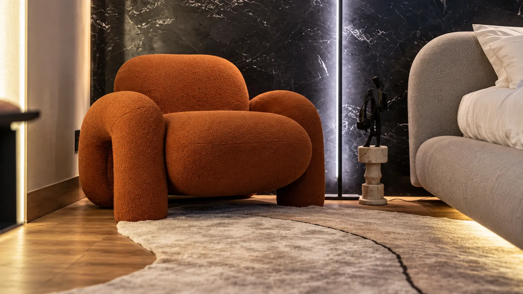 Poltrona bouclé terracota ao lado da cama, com parede de pedra iluminada